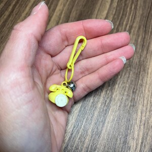 May include: A yellow telephone charm with a silver bell and a yellow clip. The miniature phone has a white dial and is attached to a yellow clip, suitable for a keychain or zipper pull.