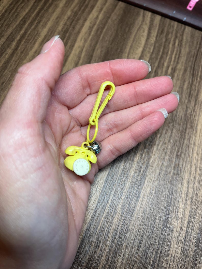 May include: A yellow telephone charm with a silver bell and a yellow clip. The miniature phone has a white dial and is attached to a yellow clip, suitable for a keychain or zipper pull.