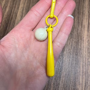 May include: A yellow baseball bat keychain with a white baseball charm. The keychain has a yellow clasp and a small ring. The baseball charm is off-white. The keychain is held in a hand.