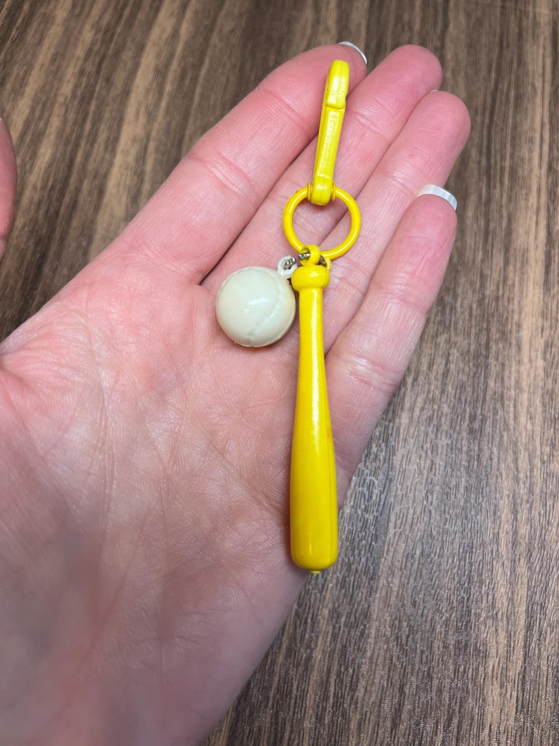 May include: A yellow baseball bat keychain with a white baseball charm. The keychain has a yellow clasp and a small ring. The baseball charm is off-white. The keychain is held in a hand.