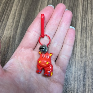 May include: A red hippo keychain with a silver bell and a red clasp. The hippo has yellow ears and blue and yellow accents on its feet. The keychain is attached to a red ring.