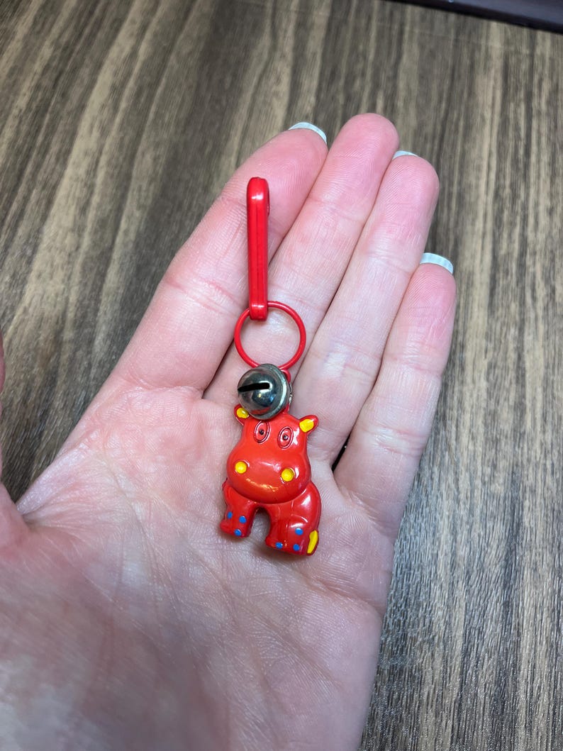 May include: A red hippo keychain with a silver bell and a red clasp. The hippo has yellow ears and blue and yellow accents on its feet. The keychain is attached to a red ring.