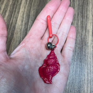 May include: A red seashell charm with a silver bead and a red clasp. The charm is attached to a red ring. The seashell has a textured surface and is held in a hand.