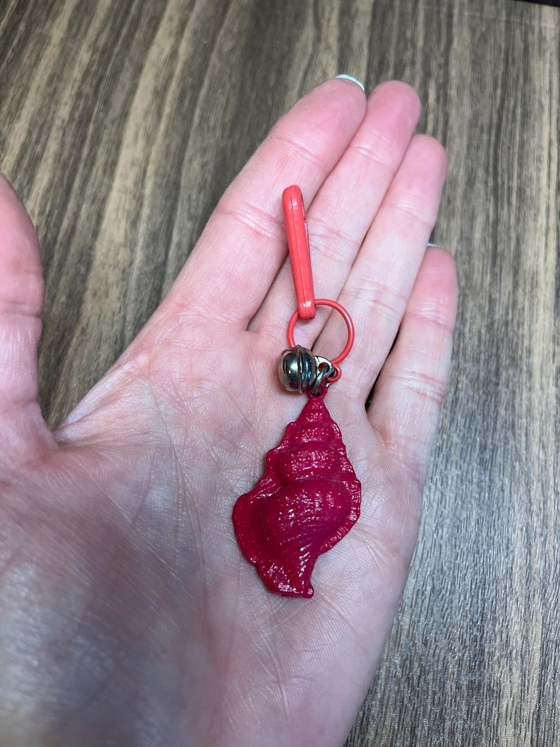 May include: A red seashell charm with a silver bead and a red clasp. The charm is attached to a red ring. The seashell has a textured surface and is held in a hand.