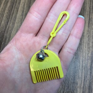 May include: A yellow plastic comb-shaped keychain with a small silver bell attached. The keychain has a yellow clip for easy attachment. The comb has vertical teeth and a rounded top.