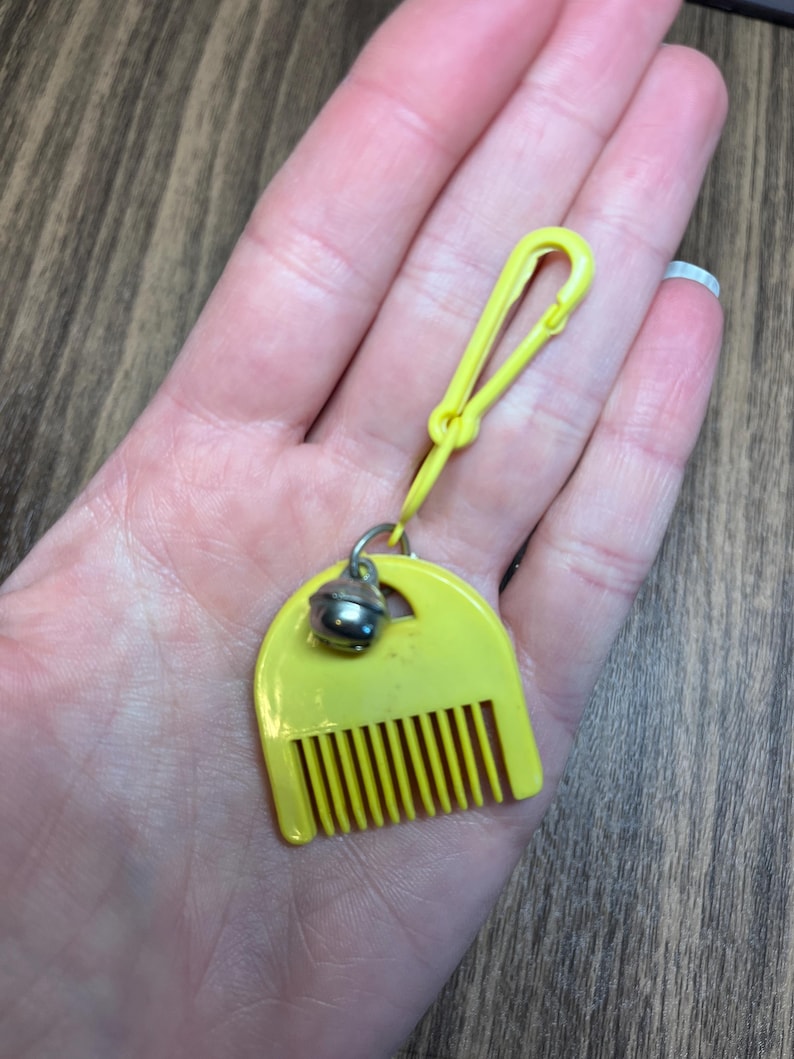 May include: A yellow plastic comb-shaped keychain with a small silver bell attached. The keychain has a yellow clip for easy attachment. The comb has vertical teeth and a rounded top.
