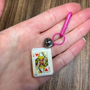 May include: Miniature playing card featuring the Queen of Hearts, paired with a silver bell charm and a pink clasp. The card has a white border and a colorful design. The bell is attached to a pink ring and clasp.