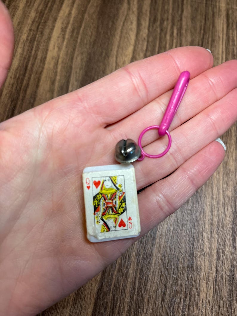 May include: Miniature playing card featuring the Queen of Hearts, paired with a silver bell charm and a pink clasp. The card has a white border and a colorful design. The bell is attached to a pink ring and clasp.