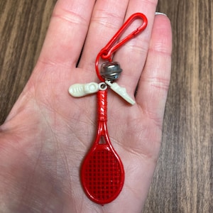 May include: A red plastic tennis racket keychain with a red clip, small silver bell, and two white plastic shoe charms. The keychain is held in a hand.