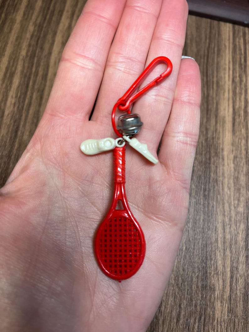 May include: A red plastic tennis racket keychain with a red clip, small silver bell, and two white plastic shoe charms. The keychain is held in a hand.