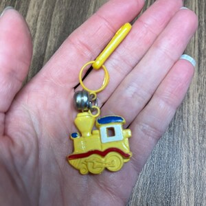 May include: A yellow train-shaped keychain with red and blue accents. The keychain has a silver bell and a yellow clip. The train has a white window and red wheels. The keychain is a fun accessory.