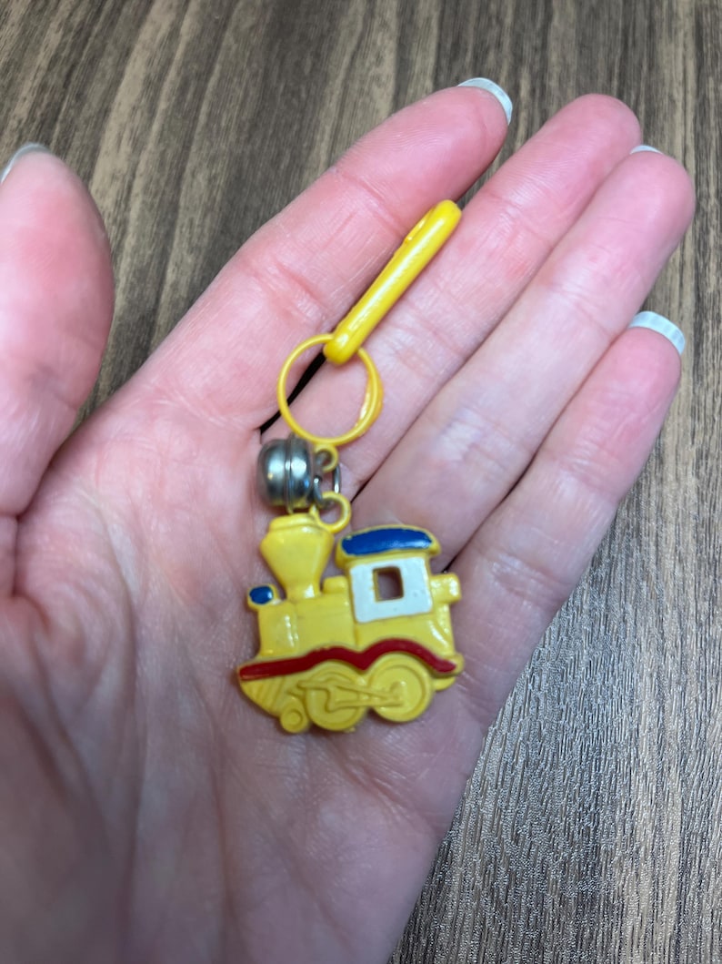 May include: A yellow train-shaped keychain with red and blue accents. The keychain has a silver bell and a yellow clip. The train has a white window and red wheels. The keychain is a fun accessory.