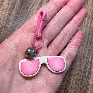 May include: A pink and white sunglasses keychain with a silver bell charm. The sunglasses have pink lenses and a white frame. The keychain includes a pink clasp and a small metal ring.