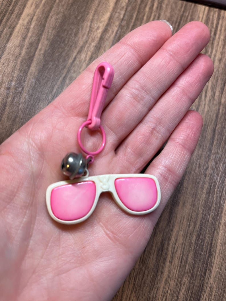May include: A pink and white sunglasses keychain with a silver bell charm. The sunglasses have pink lenses and a white frame. The keychain includes a pink clasp and a small metal ring.