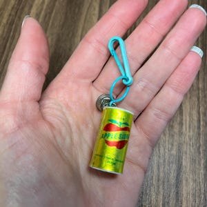 May include: A miniature, yellow Apple Cider can keychain with a red apple graphic and teal clasp. The keychain is attached to a small silver ring. The can has the words "APPLE CIDER" printed on it.