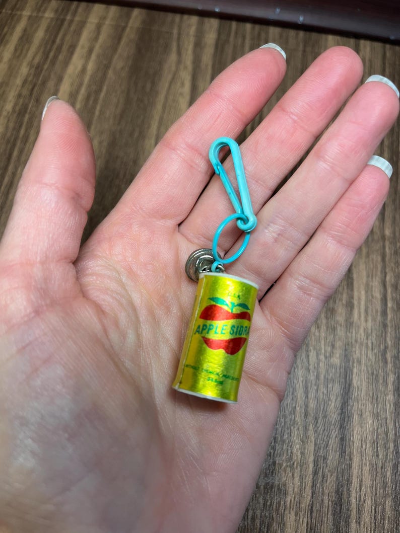 May include: A miniature, yellow Apple Cider can keychain with a red apple graphic and teal clasp. The keychain is attached to a small silver ring. The can has the words "APPLE CIDER" printed on it.