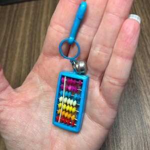 May include: A miniature abacus keychain with a bright blue frame and a matching clasp. The abacus features colorful beads in rows of red, orange, yellow, white, pink, and purple. A small silver bell hangs from the top.