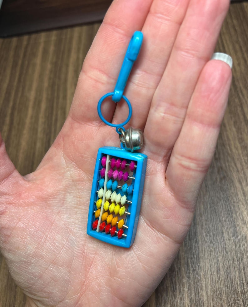 May include: A miniature abacus keychain with a bright blue frame and a matching clasp. The abacus features colorful beads in rows of red, orange, yellow, white, pink, and purple. A small silver bell hangs from the top.