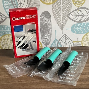 May include: Three gardening scoops with black scoop heads and teal handles, packaged in clear plastic. A red box with the Guzzini logo and the word "GREENTIME" is visible in the background.