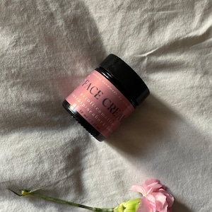 Könnte beinhalten: Ein schwarzes Gesichtscrème-Glas mit einem rosa Etikett und dem Text "FACE CREAM". Das Glas befindet sich neben einer rosa Nelkenblume auf einer weißen Stoffoberfläche. Die Creme enthält Neroli, Kamelie und Sanddorn und ist 30 ml.