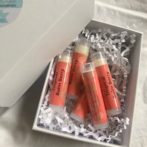May include: A white box with a lid, containing four lip balm tubes. The tubes are coral with the words "Tropical Balm Berry" printed on them. The box is filled with white paper shreds.