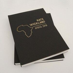 以下が含まれることがあります： 2冊の黒いノートが重ねられています。上のノートには「KATE WICKLAND AFRICA 2018」と金色の文字で書かれており、アフリカ大陸の金色の輪郭があります。