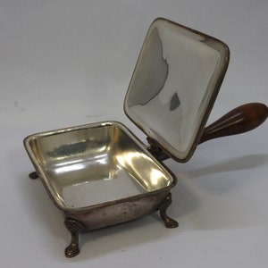 Puede incluir: Plato de servir plateado con un mango de madera y una tapa con bisagras. El plato tiene cuatro patas y es de forma rectangular.