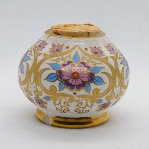 Può includere: Un piccolo vaso di ceramica bianca con tappo in sughero. Il vaso è decorato con motivi floreali dorati, viola e blu. La base e il bordo sono rifiniti in oro. Una scheggiatura è visibile sulla base.