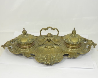 May include: An antique brass inkwell set with two inkwells and a central pen holder. The inkwells have ornate lids with a decorative design. The set is on a white background.