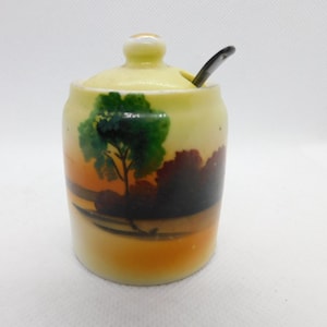 Może przedstawiać: Mały, żółty ceramiczny słoik na miód z pokrywką i małą łyżeczką. Słoik jest ozdobiony malowanym obrazkiem drzewa i zachodu słońca.