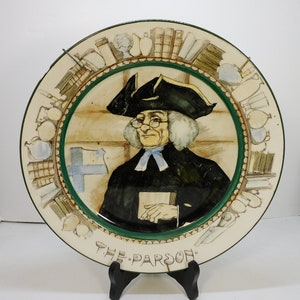May include: A vintage ceramic plate with a hand-painted image of a man in a black suit and a white wig, holding a book. The plate is decorated with a green border and a white background. The text "THE PARSON" is written at the bottom of the plate.