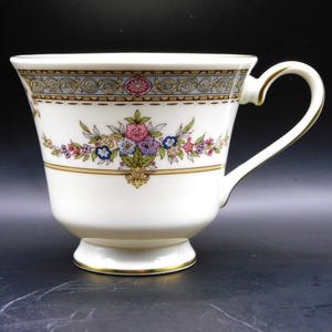 May include: A white teacup with a floral pattern in pink, blue, and purple. It has a gold rim and handle, and a decorative border with a light blue and gold design. The teacup is on a small, footed base.
