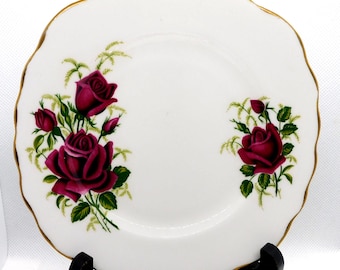 May include: A white ceramic plate with a scalloped edge trimmed in gold. The plate features two floral arrangements of deep red roses with green leaves and stems, one larger cluster on the left and a smaller single rose cluster on the right.