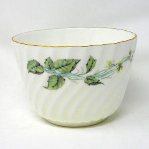 May include: White porcelain teacup with a gold rim and a delicate green leaf and vine design. The cup has a fluted exterior and a slightly irregular rim. A classic piece for tea or coffee.