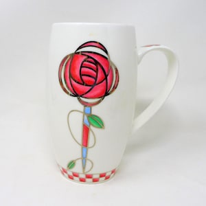 Peut inclure: Une tasse en céramique blanche ornée d'un motif de rose stylisée avec des pétales rouges, une tige bleue et rouge, et des feuilles vertes. La rose est soulignée par des lignes dorées. Un motif à damier rouge et blanc décore la base de la tasse.