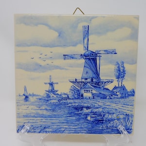 Può includere: Piastrella quadrata in ceramica con un'illustrazione blu e bianca di un paesaggio olandese. L'opera raffigura mulini a vento, acqua e un cielo nuvoloso. La scena comprende un mulino, alberi e una barca sull'acqua.