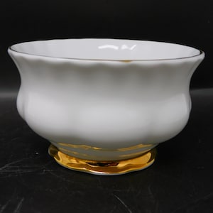 Peut inclure: Un bol en porcelaine blanc avec un bord festonné et une base dorée. Le bol a une forme arrondie et une bordure dorée autour du bord. La base est également dorée, créant un contraste avec le bol blanc.