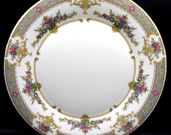 May include: A round white porcelain dinner plate featuring a decorative border with gold scrollwork, light blue geometric patterns, and colorful floral clusters in pink, purple, and green. The center of the plate is plain white.