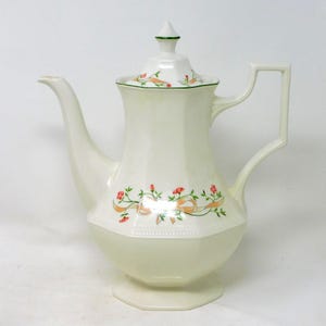Puede incluir: Tetera de cerámica color crema con diseño floral. La tetera tiene un pico largo, un asa cuadrada y una tapa con borde verde. El diseño floral incluye flores rojas y cintas naranjas.