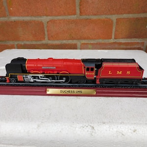 May include: A model steam locomotive painted in bright red and black with gold lettering reading LMS and 6233. The train sits on a black track mounted to a dark red base with a gold-coloured plaque that reads DUCHESS LMS in black capital letters.