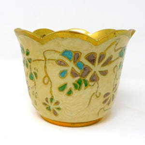 May include: A cream-coloured metal planter with a scalloped rim and a gold-toned base. The exterior features an embossed floral pattern with hand-painted details in shades of blue, green, and brown, outlined in raised gold-coloured metal lines.