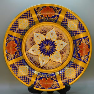 Puede incluir: Un plato decorativo con un borde dorado y diseños florales intrincados en azul, blanco y dorado. El centro del plato presenta un gran diseño en forma de estrella azul.