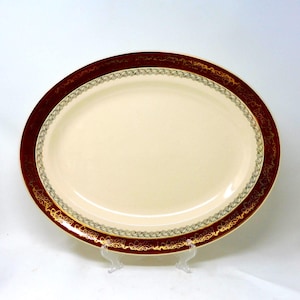 Könnte beinhalten: Eine ovale Servierplatte mit cremefarbenem Mittelteil und burgunderrotem Rand. Der Rand ist mit einem gold-grünen Dekor verziert. Die Platte wird auf einem transparenten Ständer präsentiert.