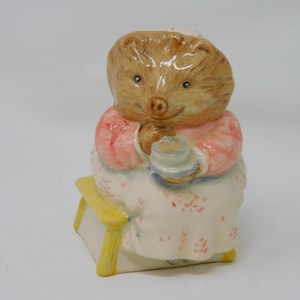 May include: A ceramic figurine of a hedgehog wearing a pink and white dress, holding a small blue object. The hedgehog is seated on a yellow bench. The figurine has a light brown head and a smiling face.