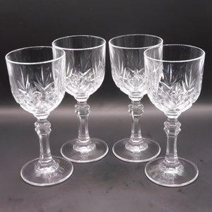 May include: Four clear crystal wine glasses with detailed starburst and vertical cut patterns. Each glass features a round base, a faceted stem, and a bowl-shaped top. The glasses are displayed on a dark surface.