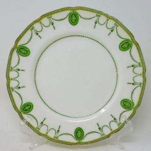 Peut inclure: Assiette en céramique blanche à bord festonné, ornée de motifs décoratifs verts et jaunes. L'assiette présente un motif répétitif d'ovales verts et de motifs en forme de guirlande, avec une bordure dorée.