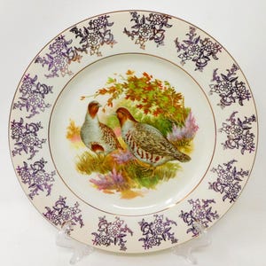 Può includere: Un piatto rotondo in ceramica con un'illustrazione centrale di due uccelli selvatici in un campo erboso con fogliame dai colori autunnali. Il bordo è decorato con un motivo floreale viola ripetuto e un sottile bordo dorato.