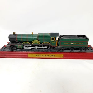 May include: A model steam locomotive painted in green with red accents and black detailing. The tender is labelled GREAT WESTERN. It sits on a red display base with a gold plaque that reads KING CLASS GWR. The train features white wheels and rods.