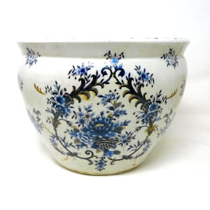 May include: A round ceramic planter with a cream-coloured base featuring a repeating pattern of dark blue and light blue floral motifs, intricate scrolling vines, and subtle gold-toned accents distributed across the exterior surface.