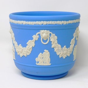 Può includere: Un vaso di ceramica blu chiaro con dettagli in rilievo bianchi. Il vaso presenta una ghirlanda decorativa, accenti a testa di leone e figure classiche. Il bordo ha un bordo floreale bianco. Un pezzo decorativo per l'arredamento della casa.